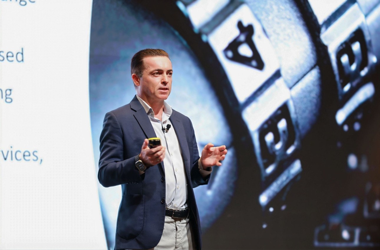 James McLeary presenting on stage with a microphone and presentation remote.
